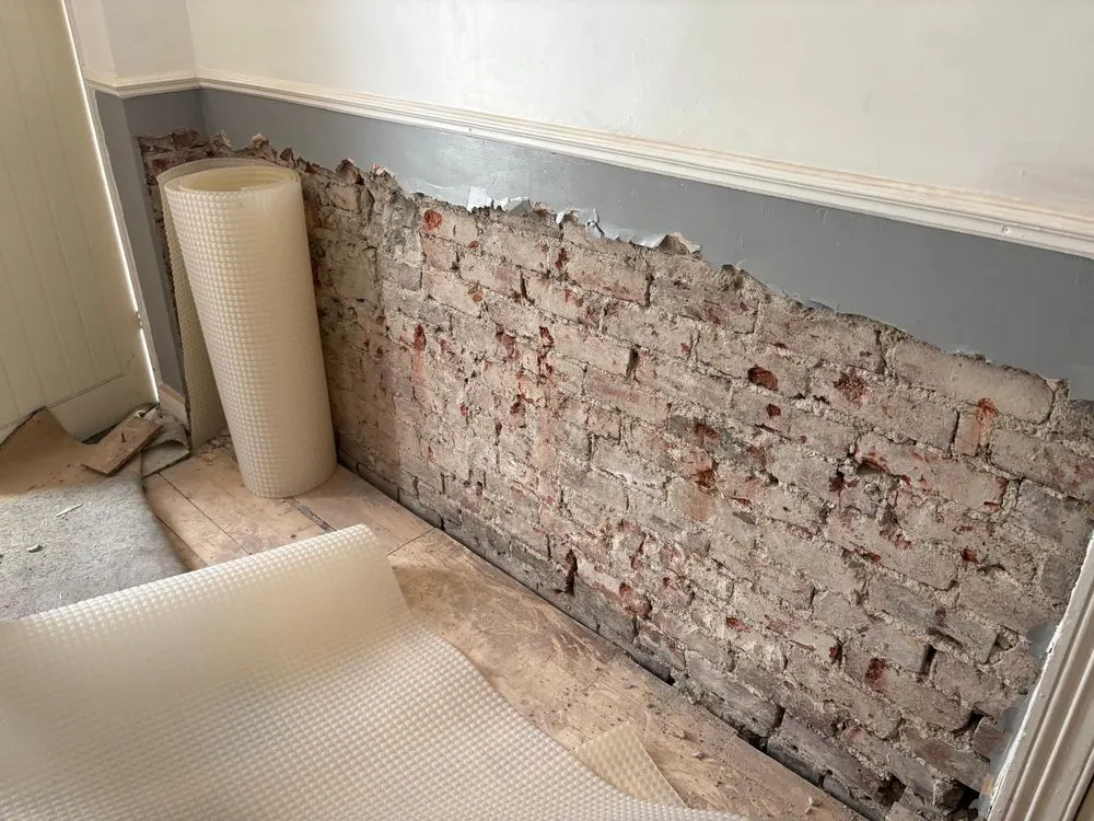 A wall stripped back to brick below the dado rail, with a roll of damp proof membrane ready to fix