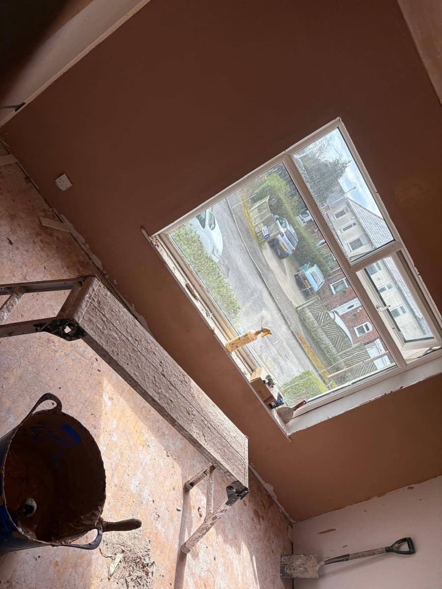 A freshly plastered wall around a window in a room being renovated, with tools and a plasterer's bucket in front
