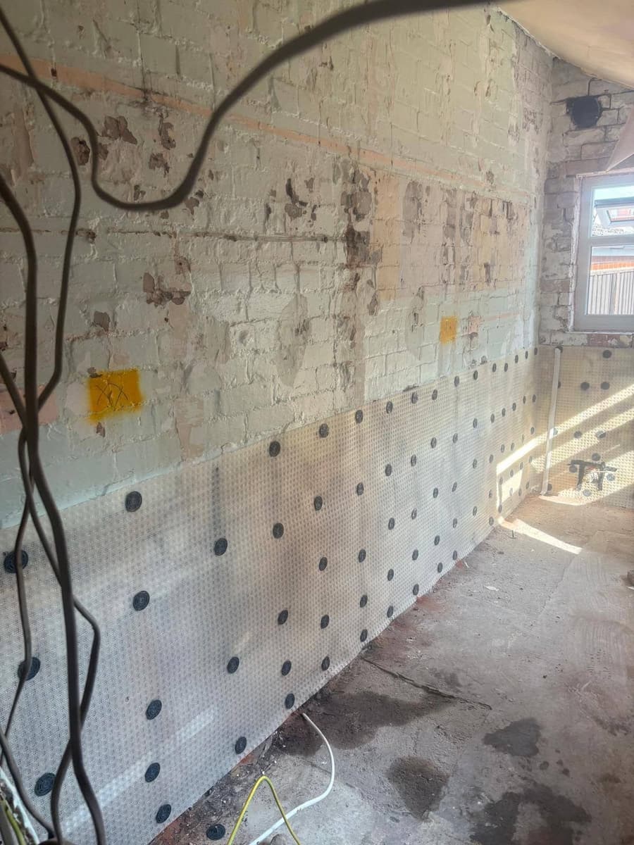 A studded membrane fixed across the lower half of a stripped brick wall, with the bare brickwork exposed above it
